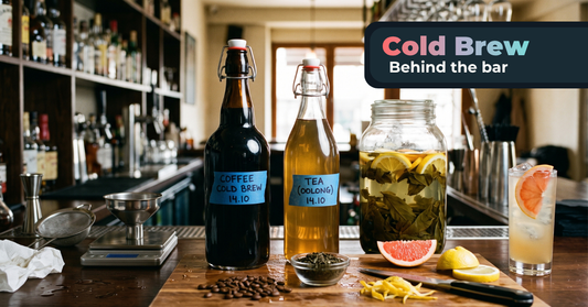 A bar counter during prep showing coffee and oolong tea cold brews in labeled swing-top bottles, a jar with steeping tea, herbs and citrus, alongside coffee beans, fresh grapefruit, lemon zest, bar tools, and a finished highball cocktail.